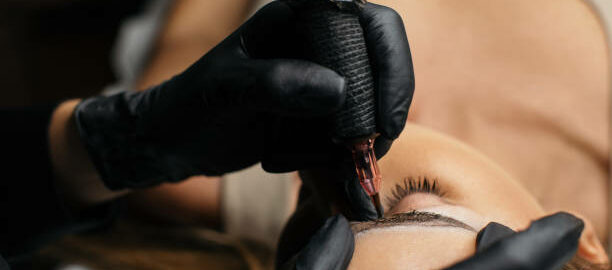 Beautician applying permanent make up microblading technique for eyebrows. She is using device and pen for special tattoo pigmentation and shaping eyebrow